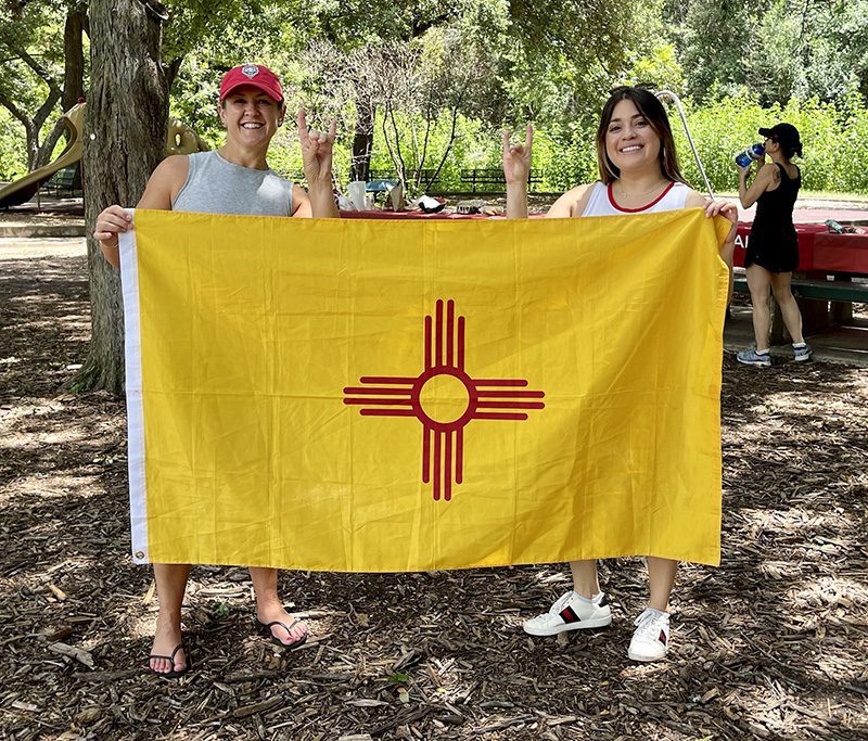 austin-chapter two women holding the NM state flag and throwing the lobo howl hand sign