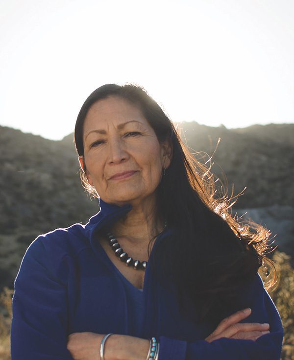 Deb Haaland wearing a blue sweater while the sun rises behind her