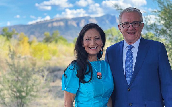 Deb Haaland in a turquoise dress with her husband, Skip Sayer, wearing a blue suit, outdoors with Sandia Peak in the background