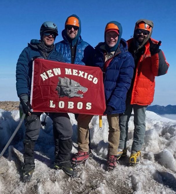 four climbers on top of a mountain with a UNM Lobos flag