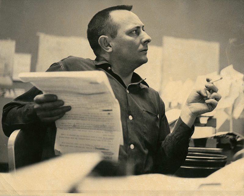 Tony Hillerman sits at desk in profile holding a large document