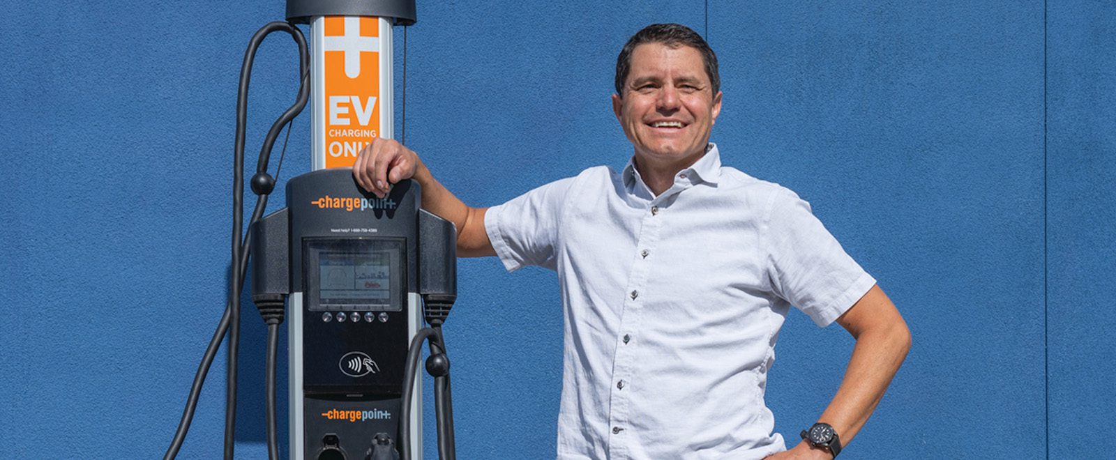 solid-state-banner Alum Doug Campbell smiling and posing with electric vehicle charging station