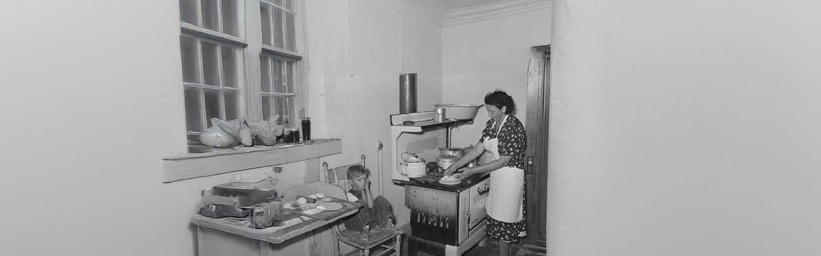 Black and white photo of woman cooking on old stove with young boy sitting in a chair looking at the person taking the photo